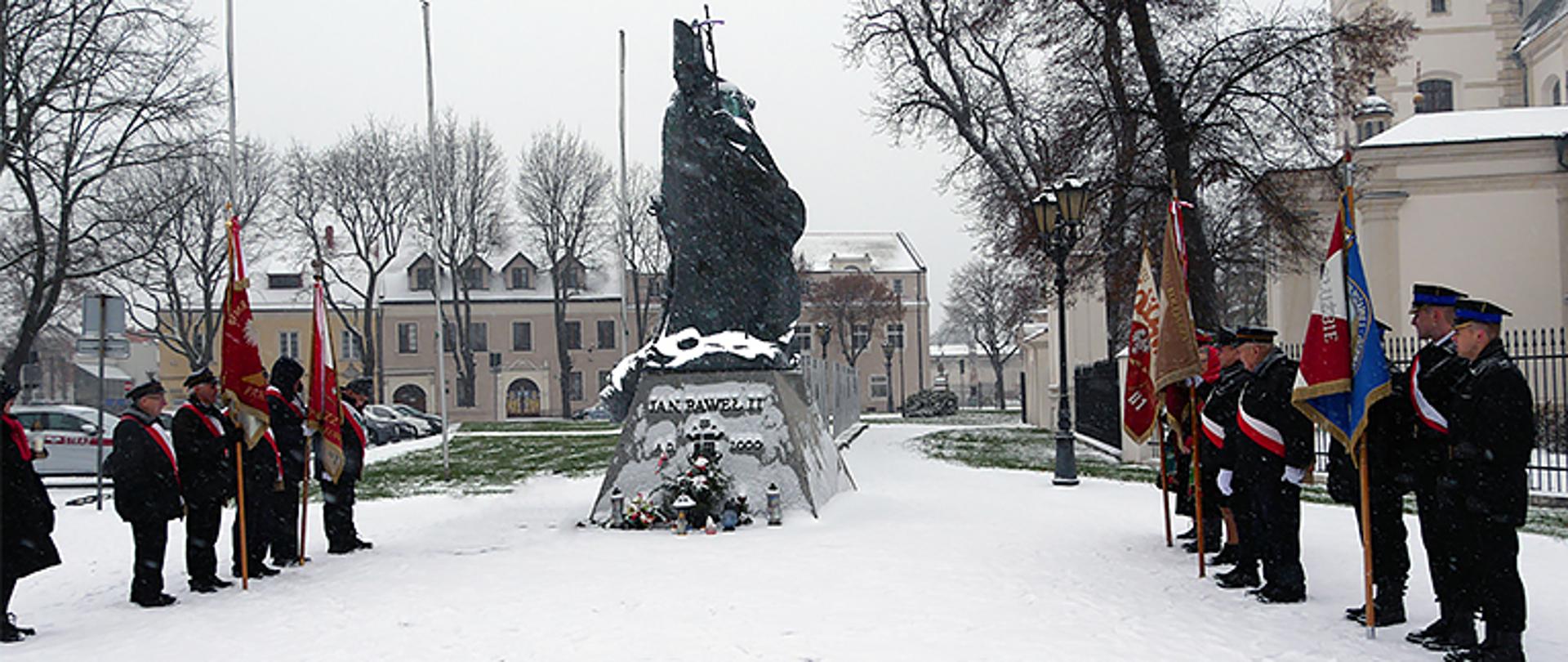Pomnik Jana Pawła II pod którym stoi pięć pocztów sztandarowych, pada śnieg