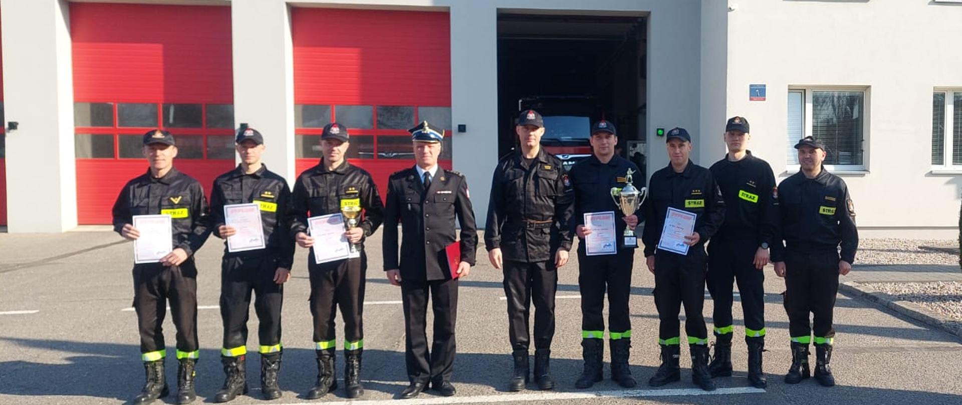 Zdjęcie przedstawia budynek koloru szarego z czerwonymi bramami. Na placu przed tym budynkiem znajduje się grupa dziewięciu strażaków w czarnych mundurach z czapkami na głowie. Pięciu strażaków trzyma w dłoni dyplom, a dwóch puchar. W środku grupy strażaków stoi komendant straży w granatowym mundurze i czapką na głowie, w dłoni trzyma czerwoną teczkę.