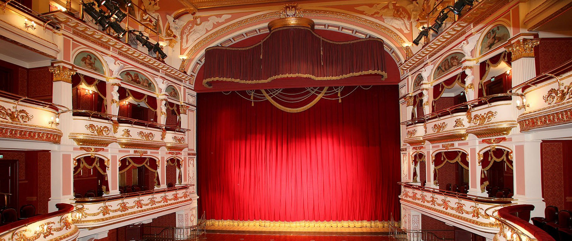 W centrum scena operowa z zasłoniętą czerwoną kurtyną , po prawej i lewej strony balkony operowe.