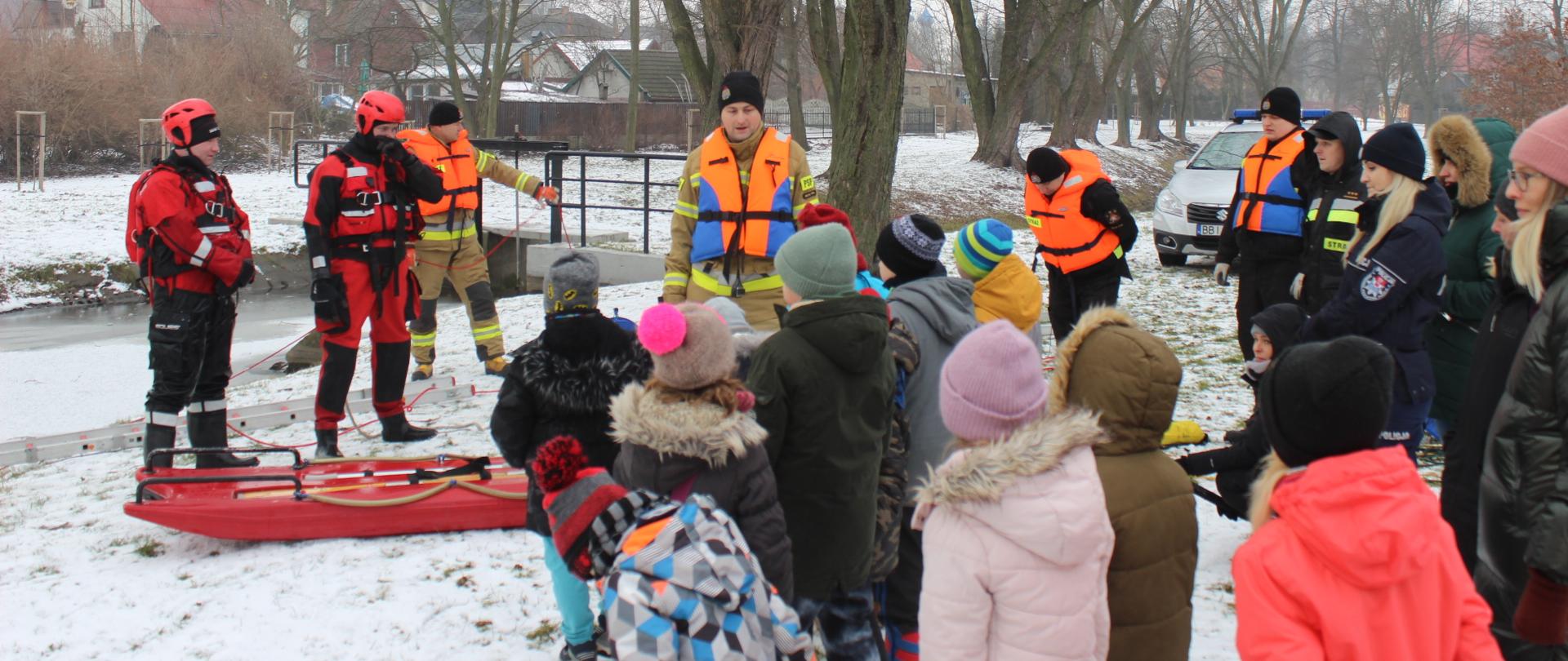 Zdjęcia przedstawia funkcjonariuszy straży pożarnej omawiających pokaz ćwiczeń lodowych dzieciom