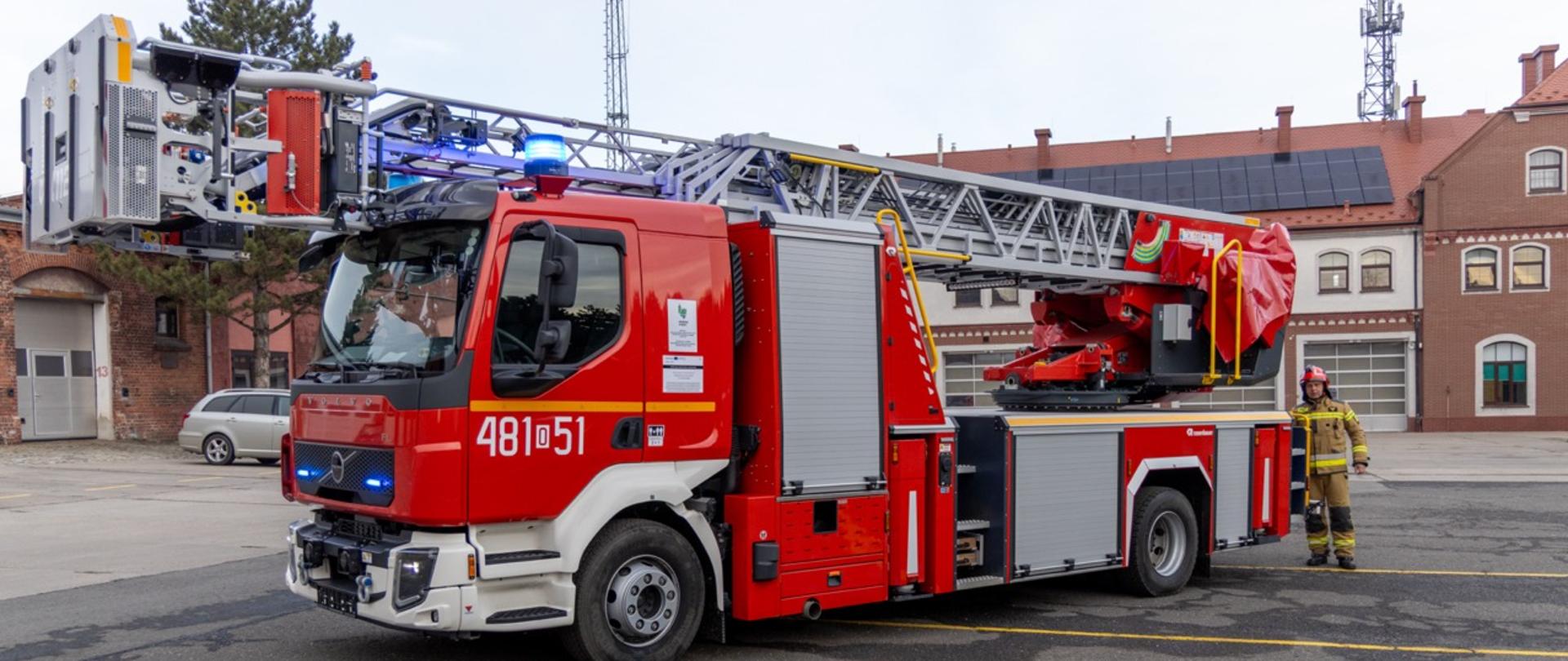 Nowa drabina mechaniczna KP PSP Prudnik o wysokości ratowniczej 30 metrów stoi rozłożona przed budynkiem jednostki. Na pierwszym planie widoczna jest drabina. W oddali strażak w pełnym umundurowaniu.