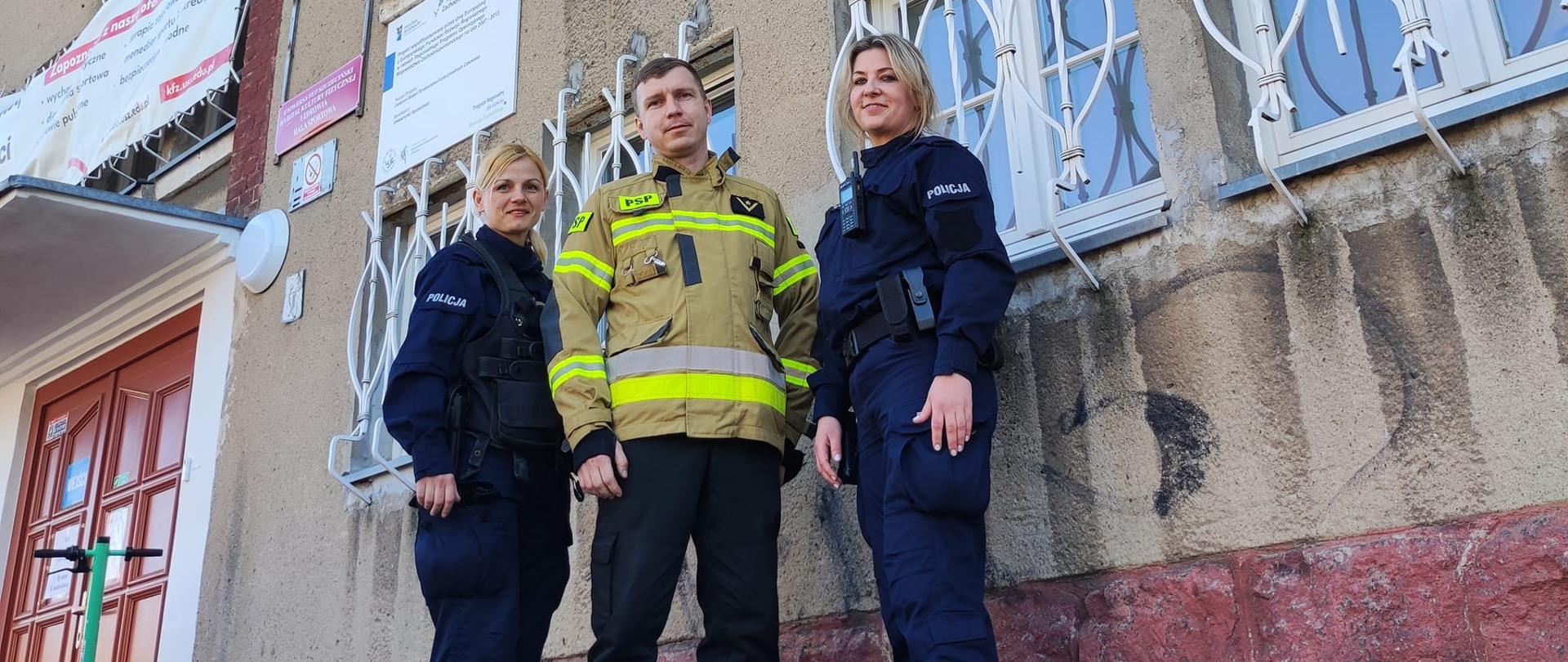 Na schodach przed budynkiem sali sportowej stoi strażak a po jego obu stronach policjantki.