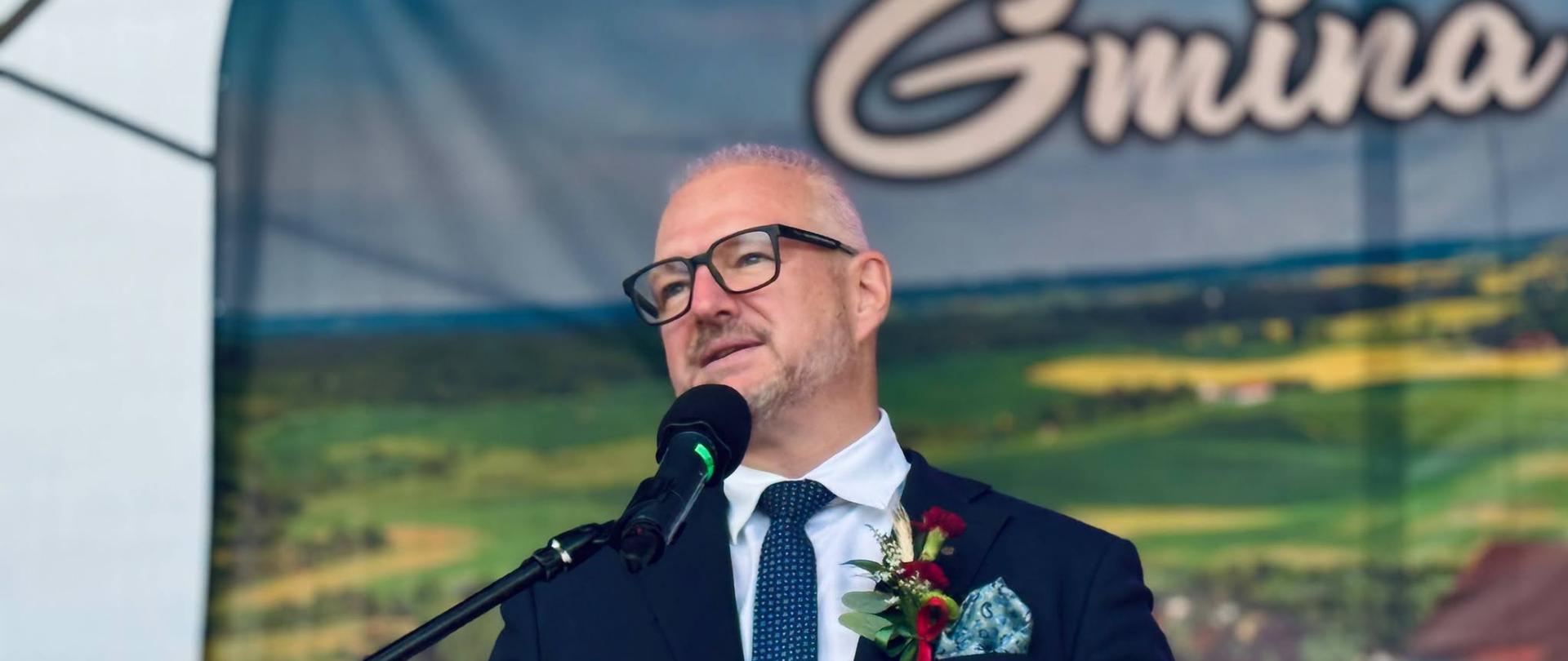 Mężczyzna w eleganckim stroju wygłasza przemówienie z podium na wydarzeniu.