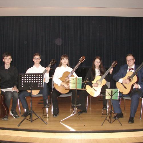 Zdjęcie przedstawia kwintet gitarowy podczas występu. Tło stanowi ciemna kotara w sali koncertowej. Widzimy pięcioro młodzieży ok 17 lat, siedzących z gitarami w rękach. Przed muzykami znajdują się pulpity a na nich nuty