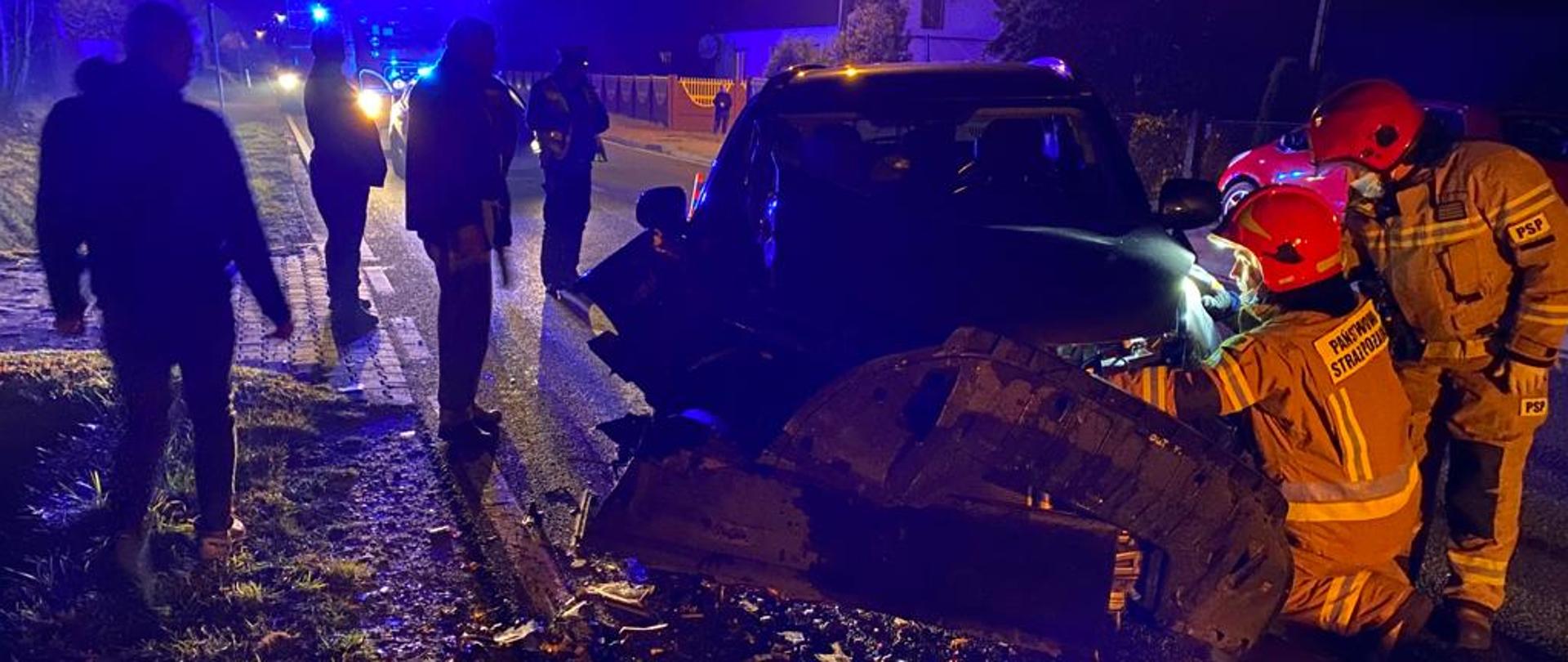 Zdjęcie robione w porze nocnej przedstawia przód uszkodzonego pojazdu osobowego. Strażacy w umundurowaniu specjalnym odłączają akumulator. W tle świadkowie zdarzenia oraz policja.