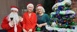 Mężczyzna w przebraniu św. Mikołaja i dwie kobiety w strojach elfów stoją przy udekorowanej choince.