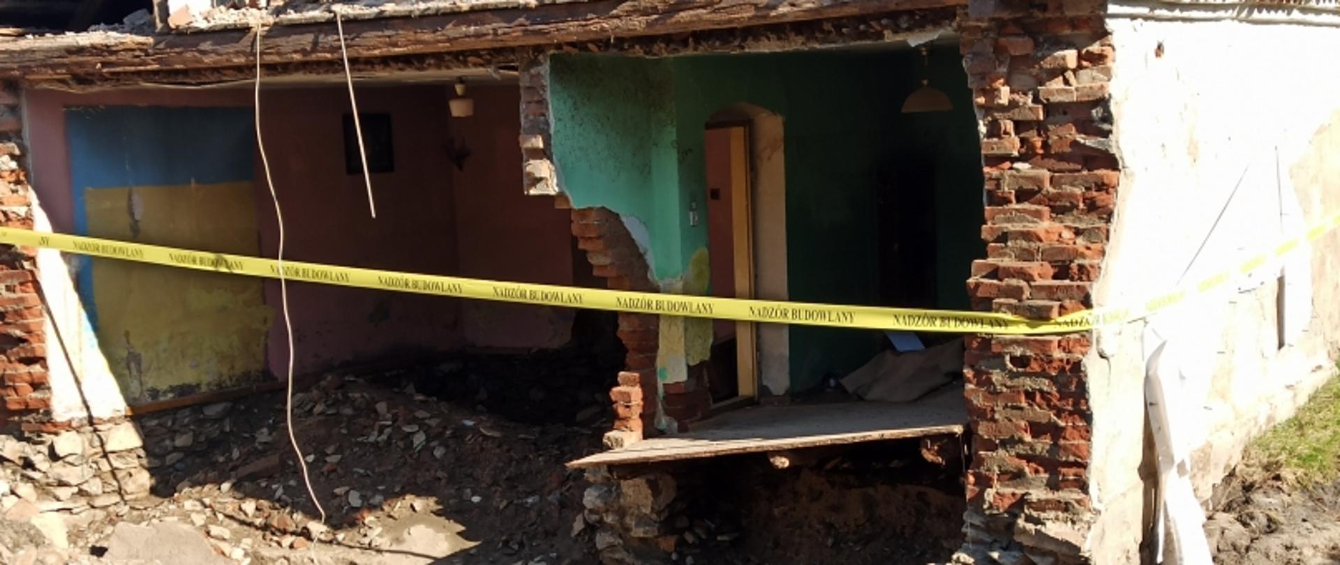 Zniszczony budynek z gruzem i taśmą ostrzegawczą; w tle dwie osoby w odzieży ochronnej.