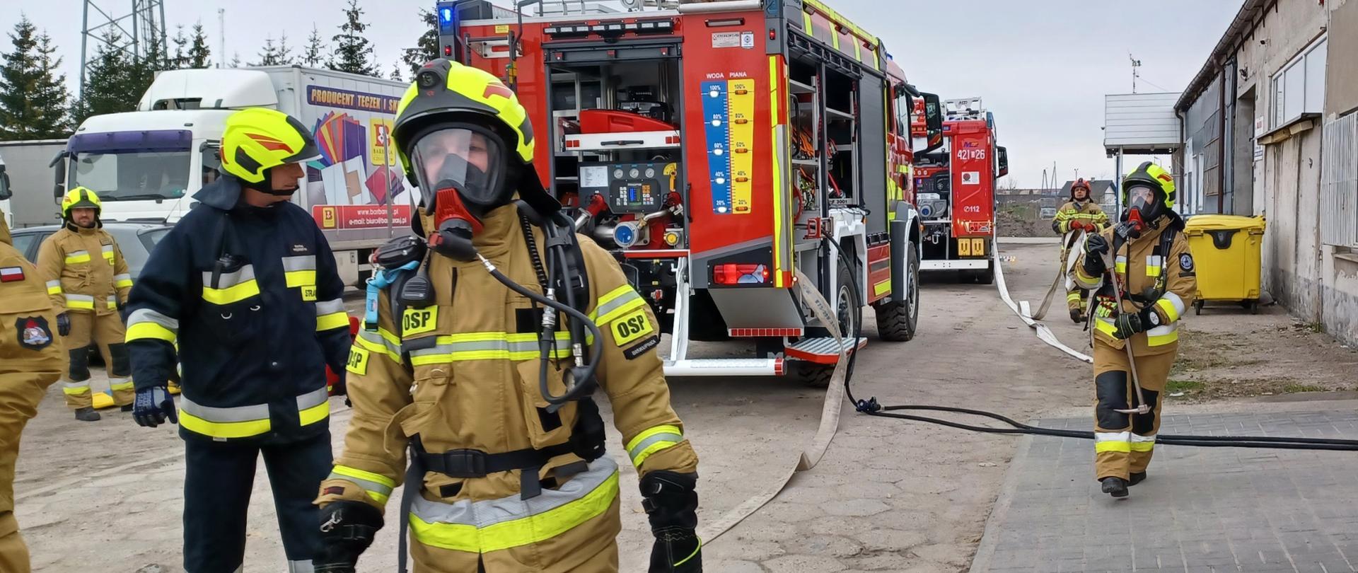 Na pierwszym planie widać strażaków w sprzęcie ochrony układu oddechowego przygotowaną do wejścia do budynku objętego pożarem oraz kierującego działaniem ratowniczym. W tle stoją samochody pożarnicze na terenie przedsiębiorstwa.