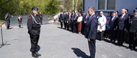 Na placu przed budynkiem strażnicy umundurowany strażak stoi naprzeciw mężczyzny w garniturze, a pozostali uczestnicy uroczystości obserwują wydarzenie.