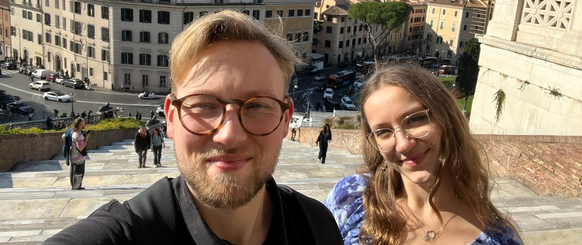 Na zdjęciu widać dwie osoby — mężczyznę i kobietę — pozujące do selfie na tle miasta Rzym. Oboje mają okulary i uśmiechają się do aparatu. W tle rozciąga się panorama miejskich budynków o jasnych elewacjach. Widać również szerokie schody, po których chodzą ludzie, oraz ruchliwą ulicę z samochodami. Pogoda jest słoneczna, niebo błękitne z kilkoma chmurkami, co nadaje zdjęciu ciepły, wakacyjny klimat.