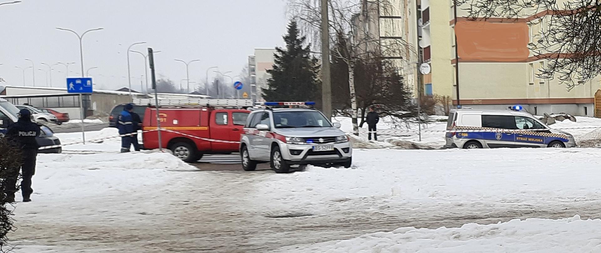 Zdjęcie obrazuje osobowy samochód strażacki osobowy samochód straży miejskiej samochód pogotowia energetycznego w lewej stronie zdjęcia stoi policjant w tle kilka osób i budynki. Aura zimowa.