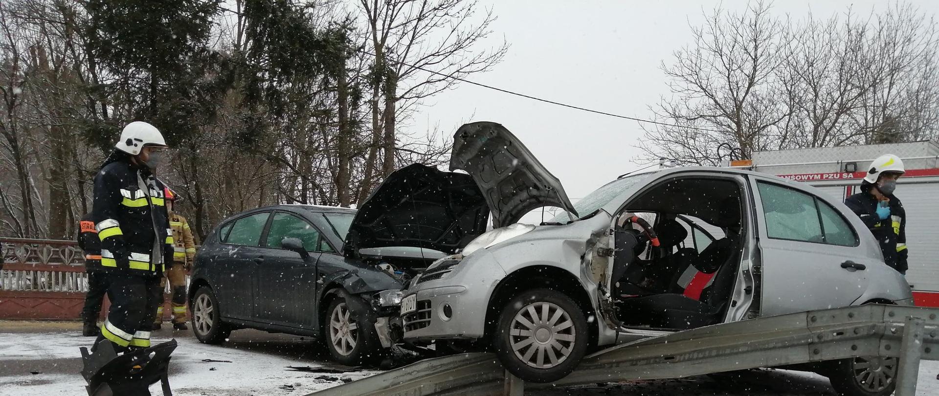 Po lewej stronie zdjęcia stoją dwaj strażacy. W środku zdjęcia widać dwa pojazdy osobowe po wypadku. Jeden z pojazdów ma usunięte przednie drzwi od strony kierowcy . Po prawej stronie widać pojazd strażacki. 