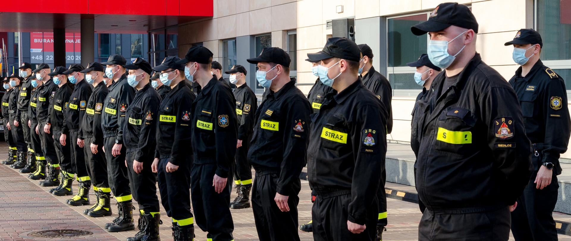 Strażacy w maseczkach ochronnych, stojący w dwuszeregu. W tle budynek KW PSP w Warszawie
