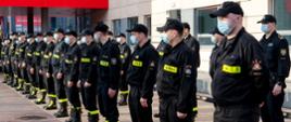 Strażacy w maseczkach ochronnych, stojący w dwuszeregu. W tle budynek KW PSP w Warszawie