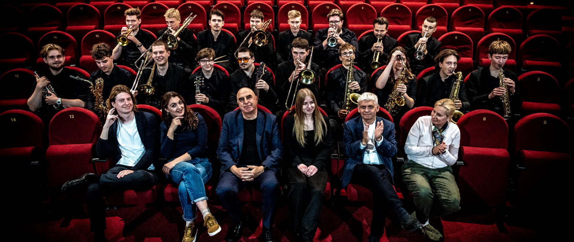 Zdjęcie przedstawia dużą grupę muzyków big-bandowych, głównie młodych mężczyzn, siedzących w trzech rzędach na tle rzędów pustych, bordowych foteli w sali koncertowej lub teatralnej. W pierwszym rzędzie na scenie siedzi pięć dorosłych osób. Mężczyzna pośrodku to prawdopodobnie dyrygent, prof. Grzegorz Nagórski. Górne rzędy zajmują instrumentaliści (saksofoniści, trębacze, puzoniści) trzymający swoje instrumenty. Oświetlenie jest dramatyczne, skupione na grupie, z ciemnym, zgaszonym tłem.