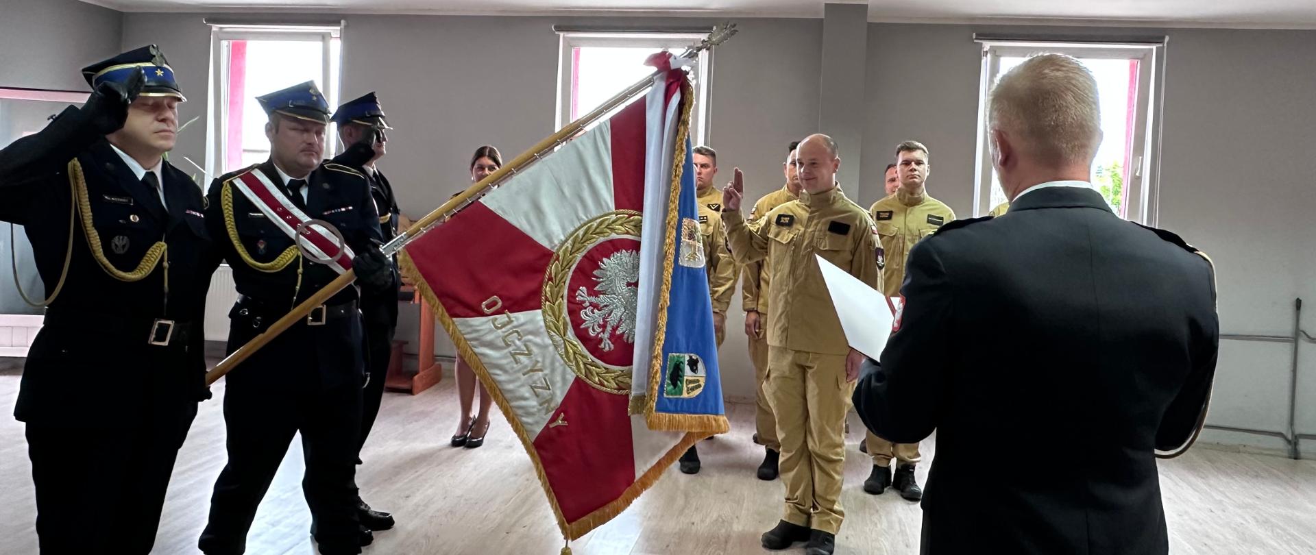 Na zdjęciu znajduje się poczet sztandarowy, Komendant oraz strażak składający ślubowanie. Na drugim planie stoją strażacy w dwuszeregu