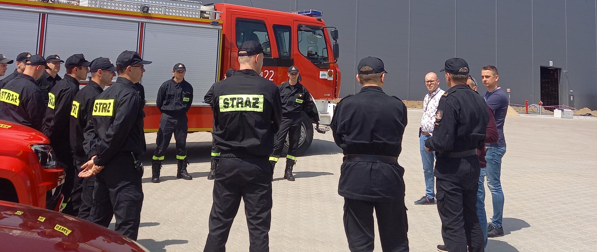 Strażacy i osoby cywilne stoją w kole. W tle samochód strażacki oraz wysoki budynek magazynowy.