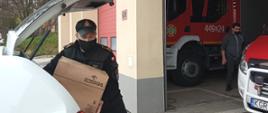 Na zdjęciu widoczny strażak z PSP w czarnym ubraniu koszarowym w czarnej czapce z daszkiem wyciągający z białego samochodu strażackiego kartony z płynem do dezynfekcji. W tle garaż jednostki OSP z samochodami pożarniczymi i strażak ochotnik w ubraniu cywilnym szara bluza i i szare spodnie.