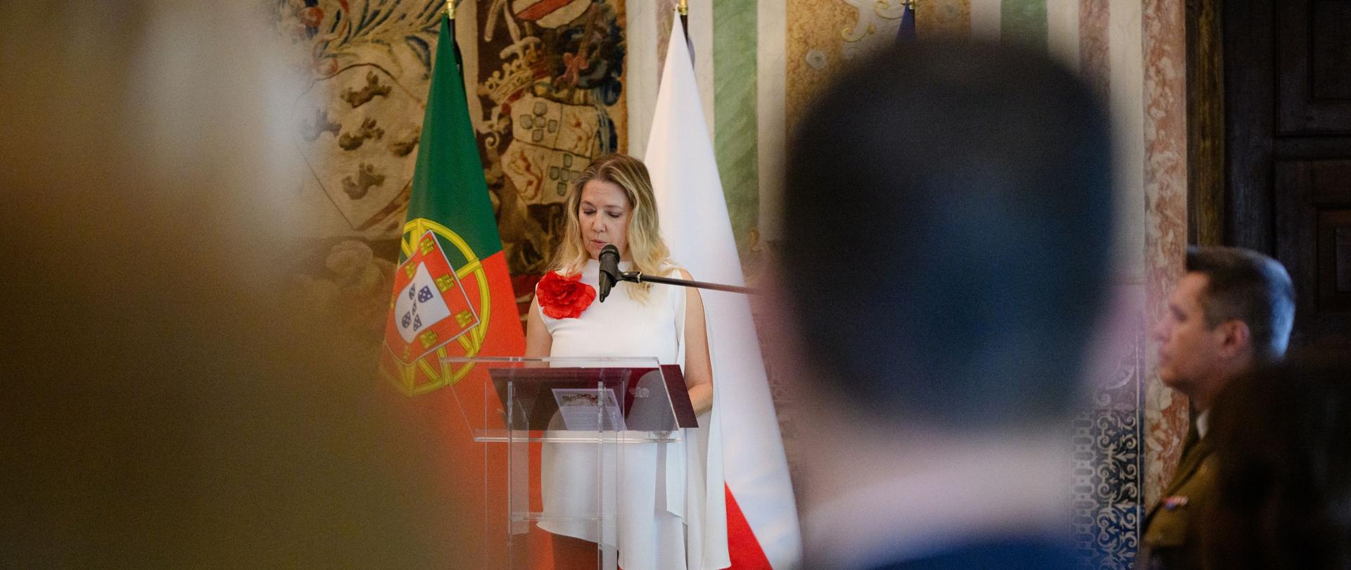 Chargée d'affaires Dorota Barys przemawia na tle flag Polski i Portugalii. Na pierwszym planie widoczne są rozmyte sylwetki uczestników wydarzenia.