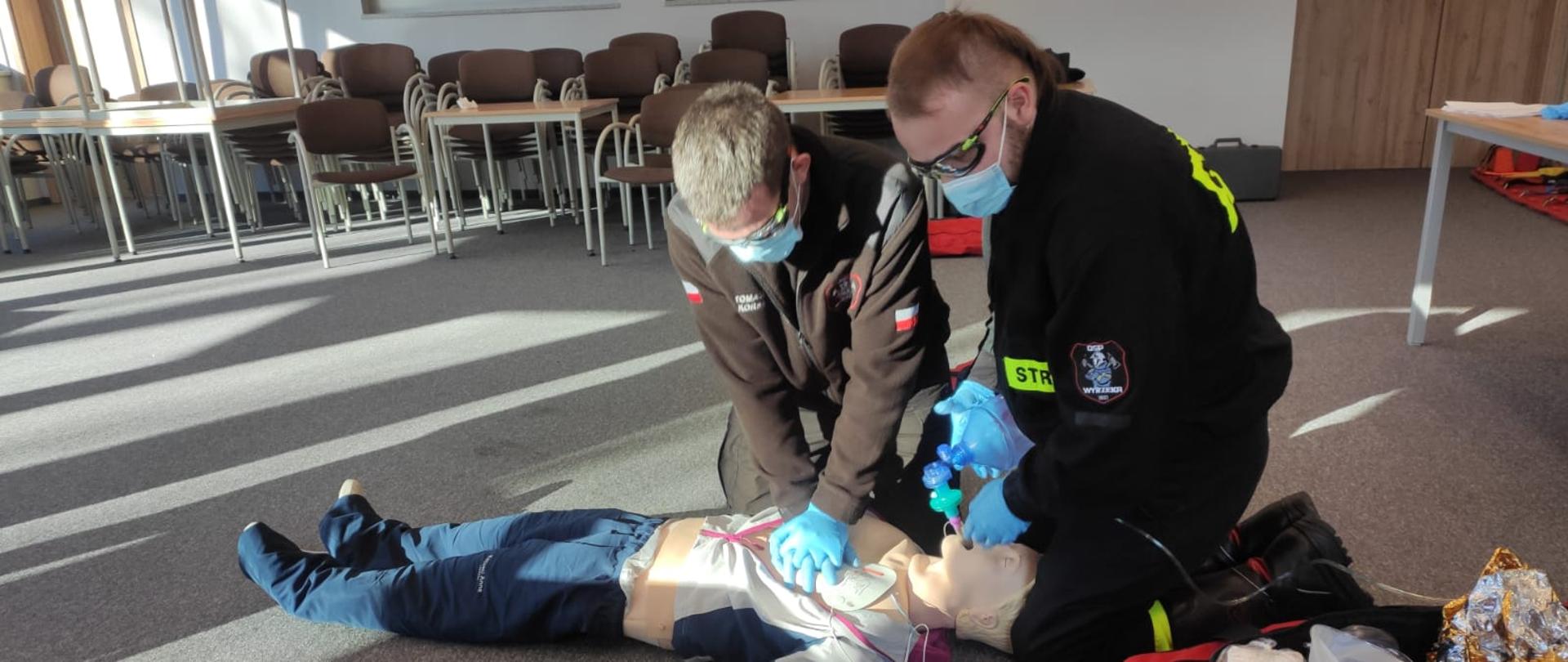 Na zdjęciu dwóch ratowników pochylonych nad fantomem i prowadzący zewnętrzny masaż serca, obok defibrylator oraz torba medyczna koloru czerwonego