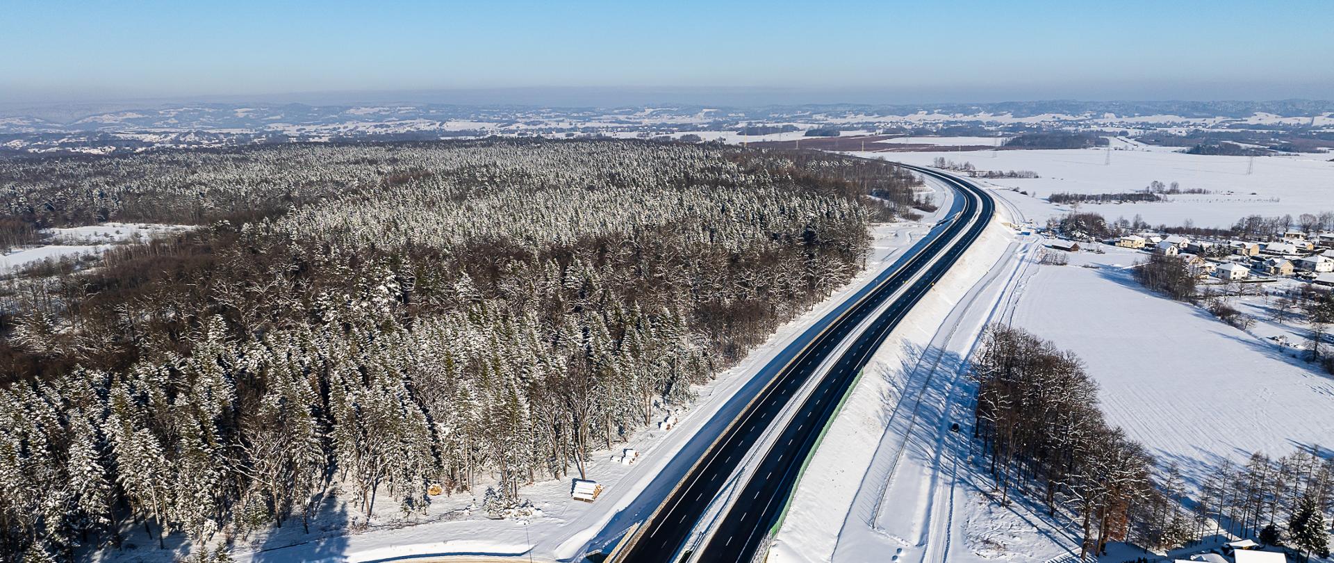 Zdjęcie wykonane z lotu ptaka przedstawia szeroką, dwupasmową drogę ekspresową biegnącą przez zimowy krajobraz. Po lewej stronie szosa sąsiaduje z gęstym lasem pokrytym grubą warstwą śniegu. Po prawej rozciągają się otwarte, białe pola z pojedynczymi zabudowaniami. Droga jest czarna i odśnieżona, a wokół dominują jasne, śnieżne przestrzenie. Na horyzoncie widać łagodne pagórki i błękitne niebo bez chmur.