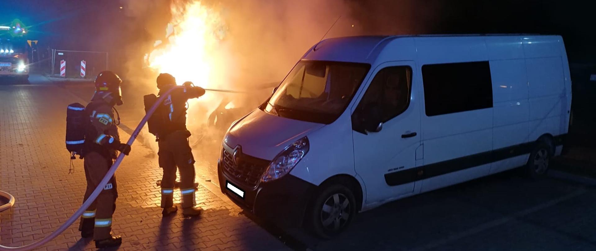 Samochód osobowy płonie na parkingu. Obok stoi samochód dostawczy. Strażacy przy pomocy węża gaszą płonący pojazd.