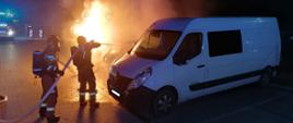 Samochód osobowy płonie na parkingu. Obok stoi samochód dostawczy. Strażacy przy pomocy węża gaszą płonący pojazd.