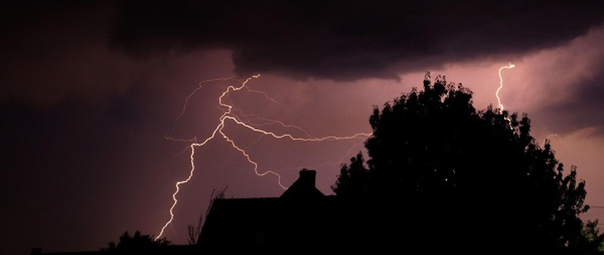 na zdjęciu widać z daleka dom a obok drzewa nocą, na niebie pioruny i czarne chmury, burza