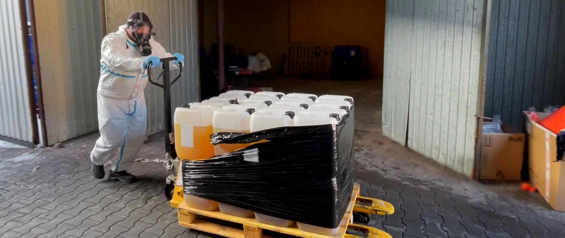 Osoba w odzieży ochronnej przesuwa wózek paletowy z owiniętą folią paletą pełną kanistrów.