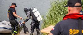 Nurek Państwowej Straży Pożarnej przygotowuje się do wejścia pod wodę. Asekuruje go strażak w kamizelce asekuracyjnej stojący na brzegu akwenu. Z boku widoczny ponton na brzegu. W tle akwen.