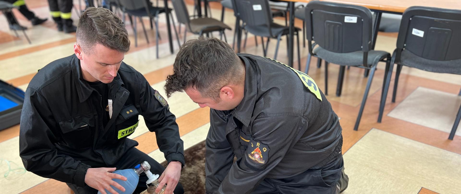 Na pierwszym zdjęciu strażacy-ratownicy wykonują symulację resuscytacji krążeniowo-oddechowej u osoby dorosłej. Jeden z ratowników wykonuje 30 uciśnięć klatki piersiowej, natomiast drugi wykonuje 2 oddechy ratownicze za pomocą worka samorozprężalnego.