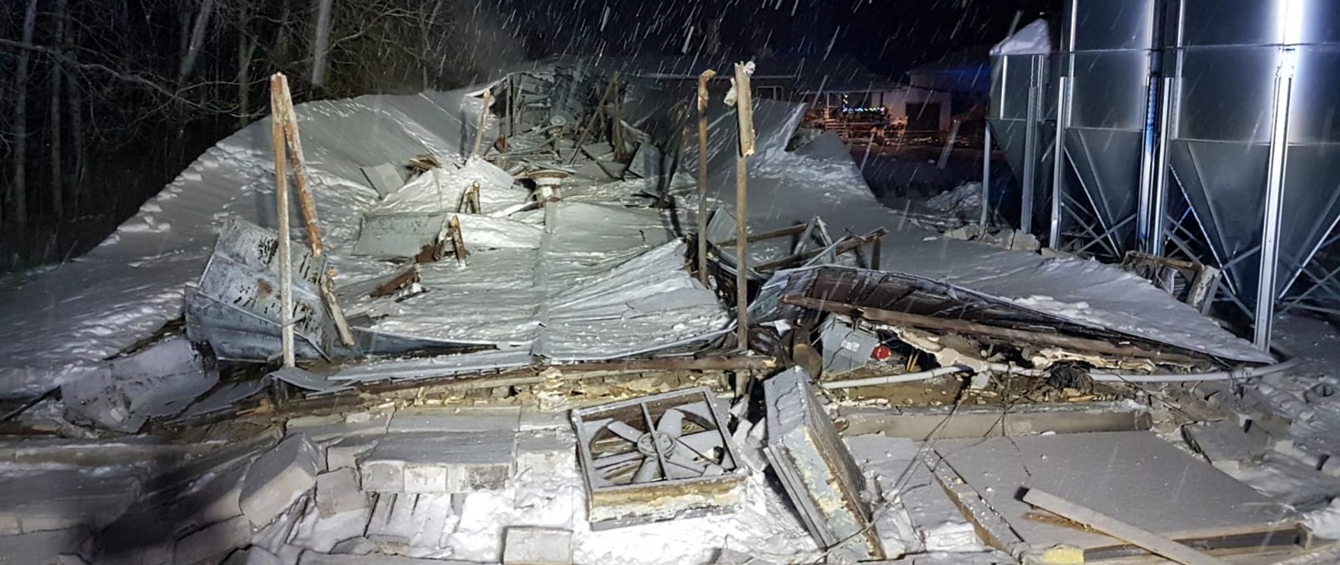 Widać cegły zawalonej ściany budynku. W dalszej części leży oberwany dach. Po prawej stronie stoją silosy na paszę.