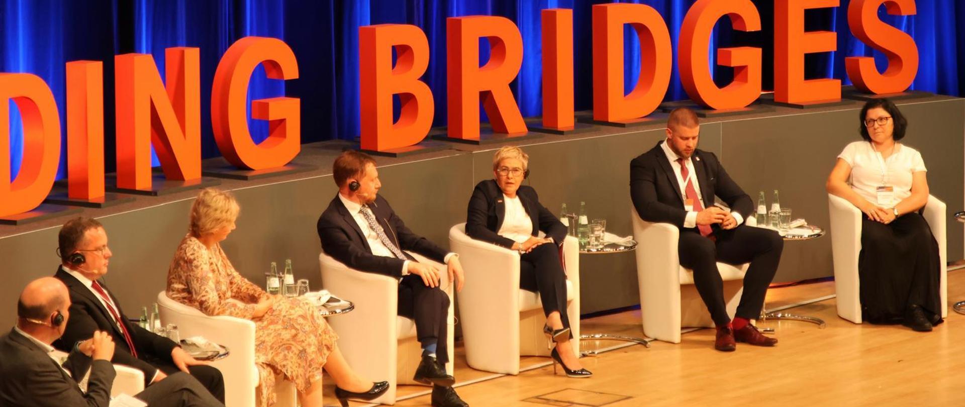 Pod ścianą z napisem Building Bridges siedzi kilka na białych fotelach osób.