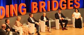Pod ścianą z napisem Building Bridges siedzi kilka na białych fotelach osób.