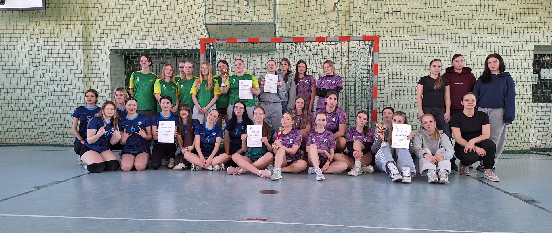 Szerokie, poziome ujęcie dużej grupy młodych kobiet pozujących na parkiecie sali gimnastycznej przed bramką do futsalu. Dziewczęta są ubrane w różne stroje sportowe (zielone, fioletowe, niebieskie koszulki). Część z nich siedzi w pierwszym rzędzie na podłodze, część stoi za nimi. Kilka zawodniczek trzyma w rękach dyplomy i małe trofea. Tło stanowi zielona siatka ochronna na ścianie i kosz do koszykówki.