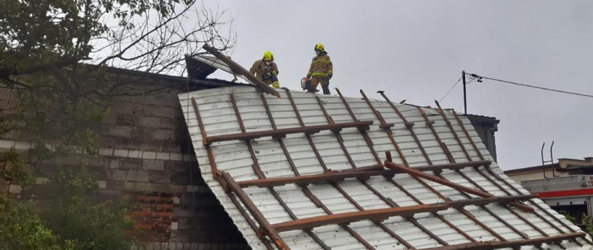 Pochmurno, zerwany dach z blachy falistej z przytwierdzonymi do niego krokwiami. Dwóch strażaków w jasnych ubraniach i żółtych hełmach stoi na części dachu. Jeden w rękach trzyma piłę. Przed budynkiem stoi wysokie drzewo liściaste.