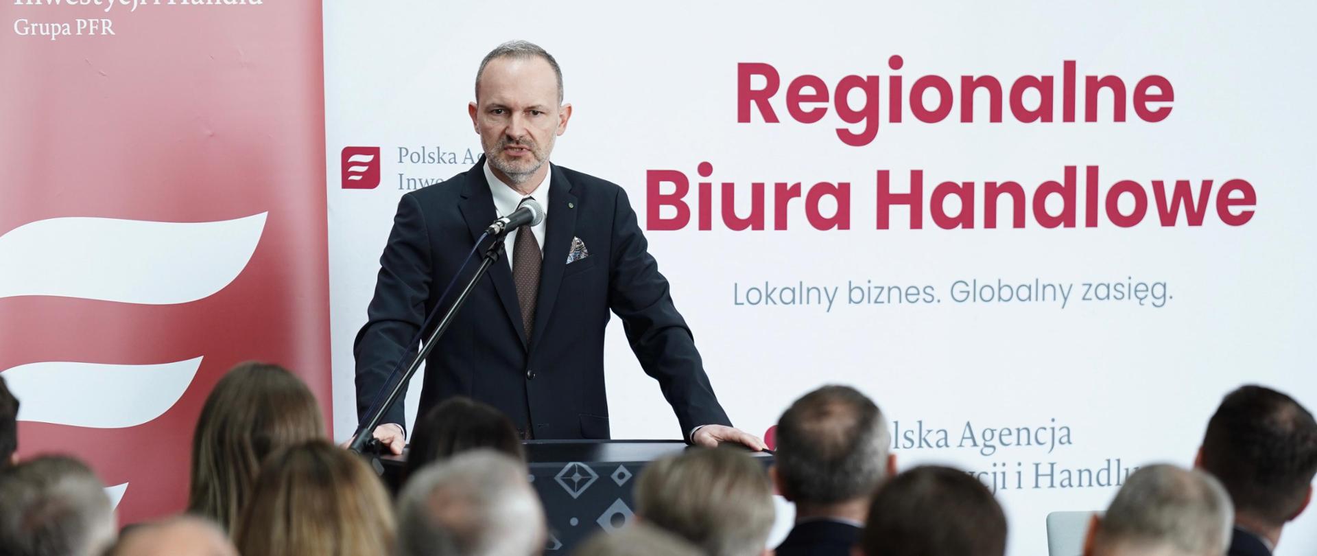 Minister Hetman przemawia przed grupą ludzi. W tle napis Regionalne biura handlowe. Lokalny biznes. Globalny zasięg. 