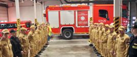 Strażacy stoją w garażu w dwóch szeregach zwróconych naprzeciwko siebie. W tle stoi zaparkowany ciężki samochód gaśniczy.