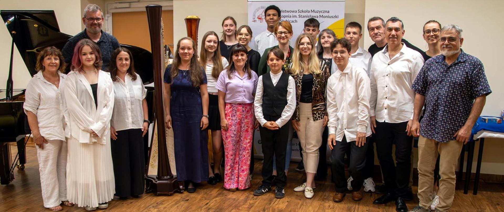 Na zdjęciu, w auli szkoły stoi grupa kilkunastu osób, dorosłych i młodzieży, Obok nich stoi fortepian, pośrodku dwie harfy, za nimi stoi baner szkolny.
