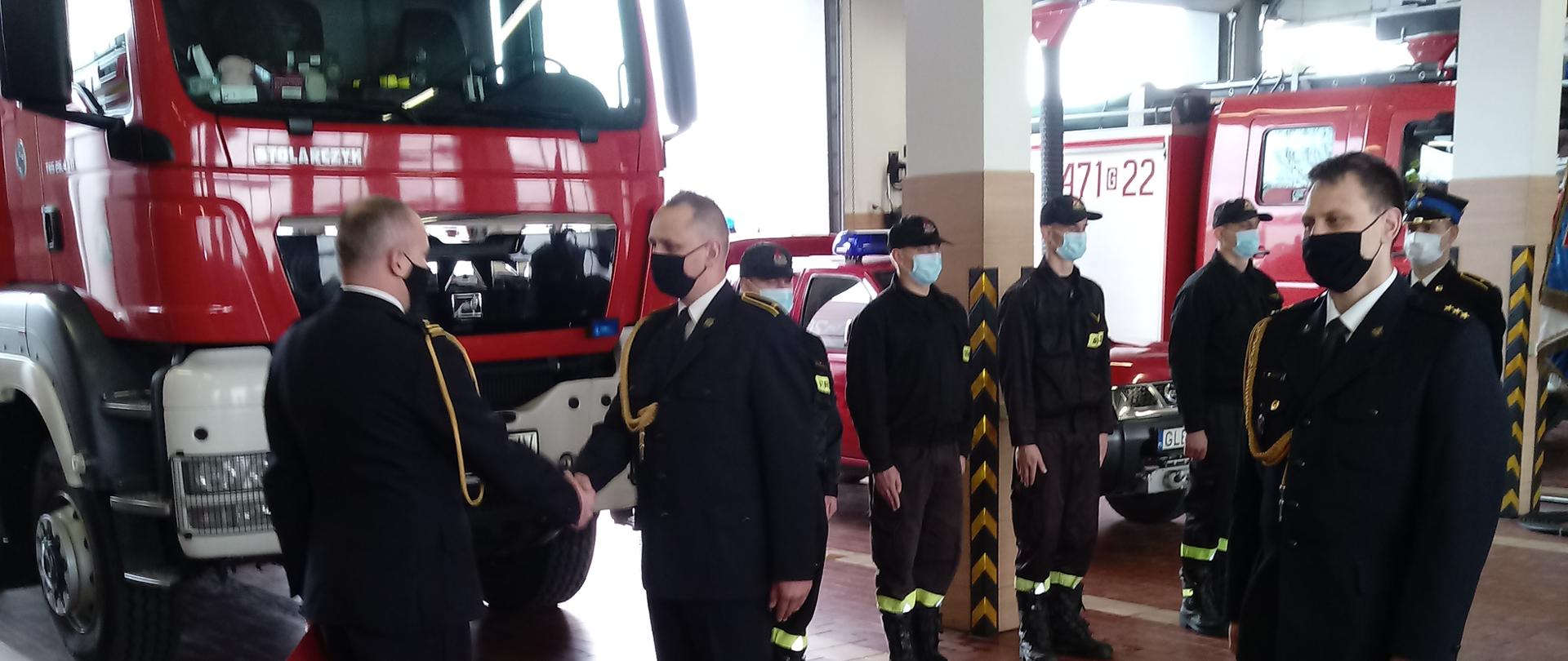 Zdjęcie przedstawia Komendanta Powiatowego Państwowej Straży Pożarnej w Lęborku wręczającego akt mianowania zastępcy dowódcy Jednostki Ratowniczo-Gaśniczej. Po prawej stronie widoczny dowódca Jednostki Ratowniczo-Gaśniczej w tle zmiana służbowa oraz pojazdy pożarnicze.