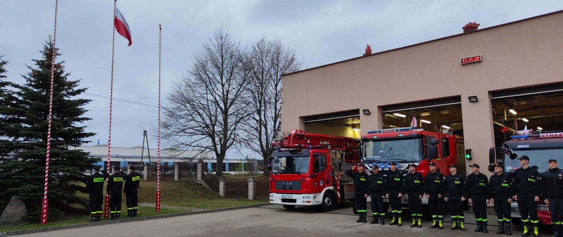 Zdjęcie przedstawia strażaków ustawionych w szyku n tle samochodów pożarniczych. Po lewej stronie strażacy wciągają flagę państwową na maszt.