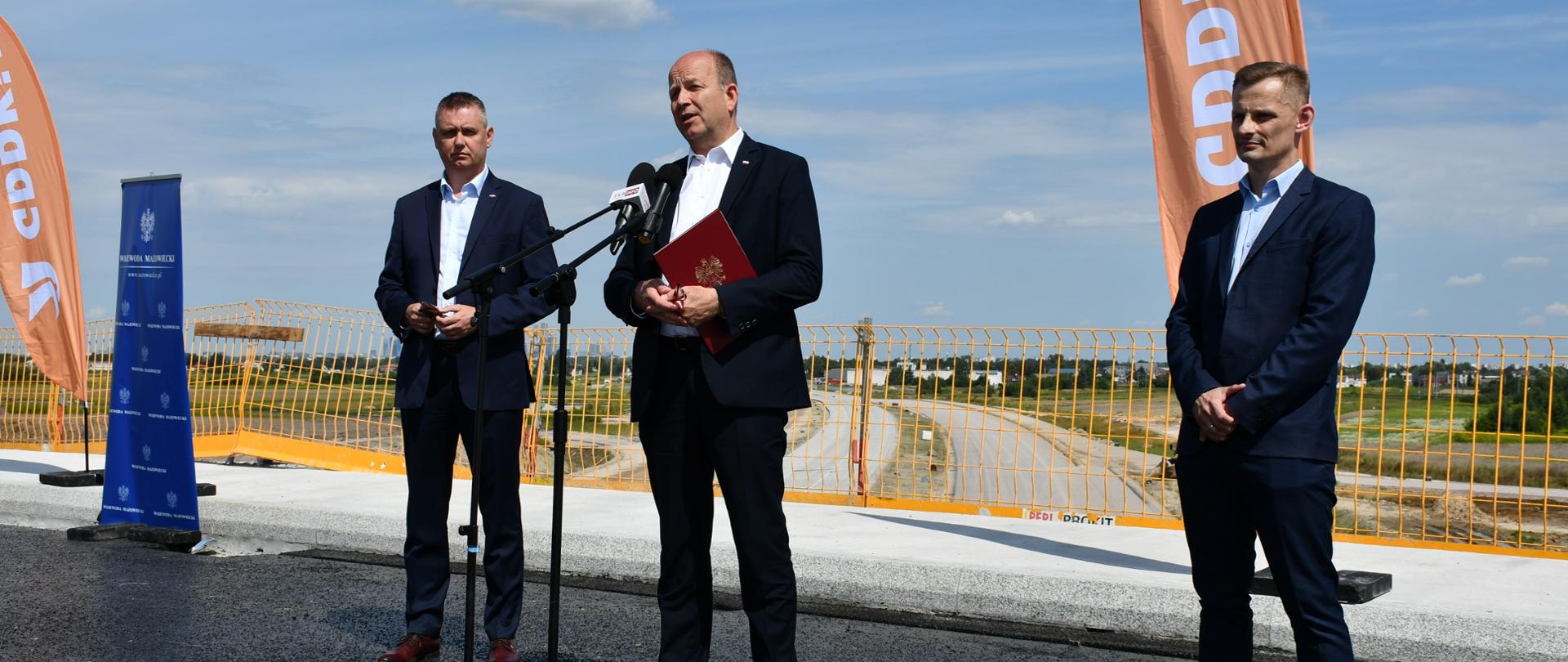 Na zdjęciu znajduje się trzech elegancko ubranych mężczyzn. Mężczyźni Ci znajdują się na wiadukcie drogowym, znajdującym się nad przygotowywaną drogą ekspresową S7 relacji Warszawa - obwodnica Grójca. W tle znajduje się fragment budowy tej drogi. 