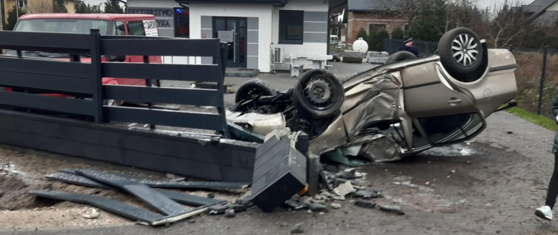 Na zdjęciu widać leżący na dachu, rozbity samochód osobowy oraz zdewastowane ogrodzenie prywatnej posesji. 