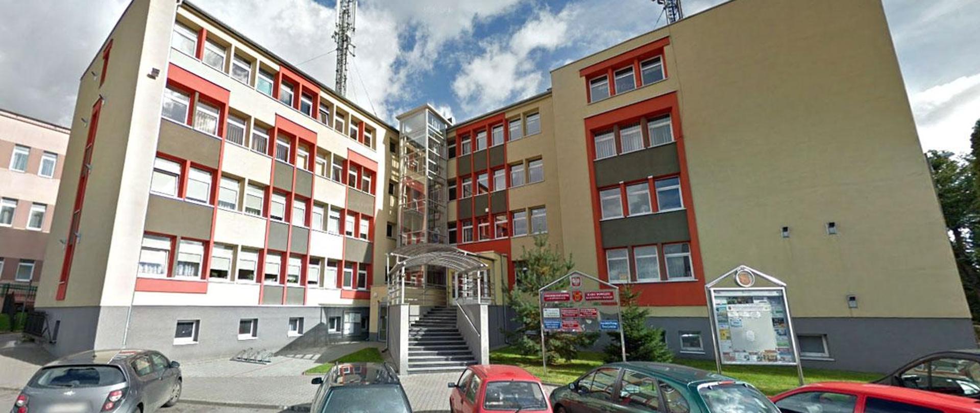 Trzypiętrowy budynek z charakterystyczną, kolorową elewacją. Przed budynkiem znajduje się niewielki parking.