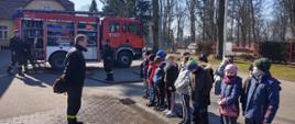 Na zdjęciu widać samochód gaśniczy, strażaka i stojącą przed nim w szeregu młodzież szkolną
