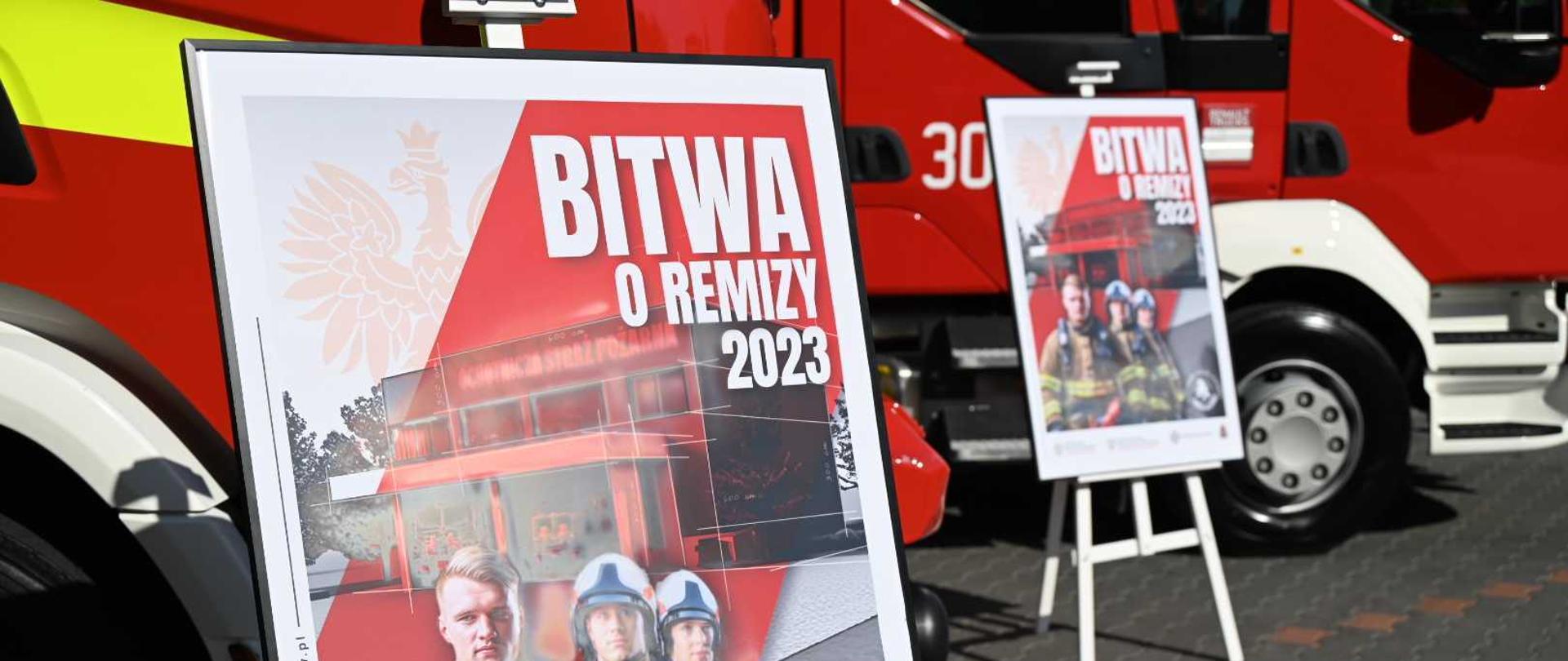 Dwa plakaty promocyjne akcji &quot;Bitwa o Remizy 2023&quot; na tle dwóch pojazdów pożarniczych