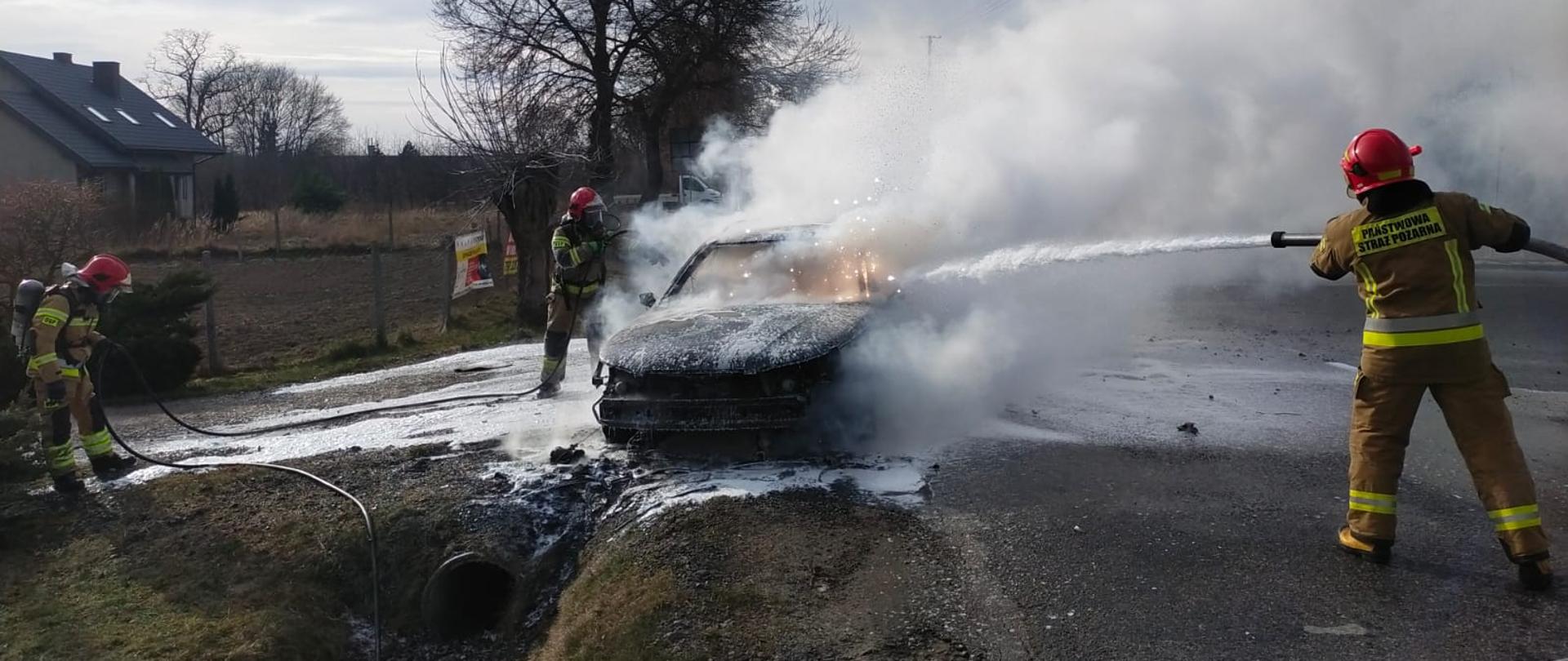 Samochód osobowy jest gaszony przez trzech strażaków w pełnym stroju strażackim. Samochód stoi na poboczu drogi asfaltowej.