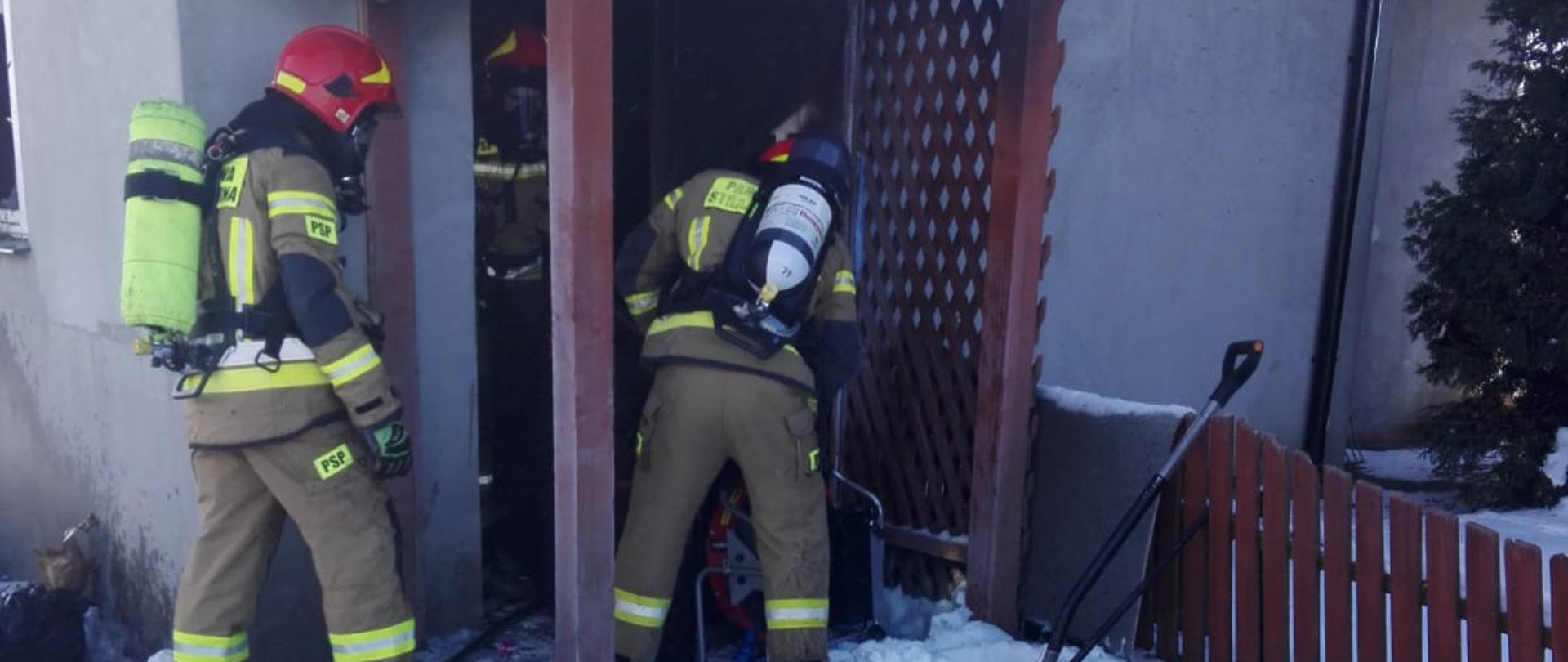 Na zdjęciu znajduje się trzech strażaków, którzy wchodzą do budynku. Przed wejściem do budynku na śniegu widoczne są leżące kartony i segregatory.
