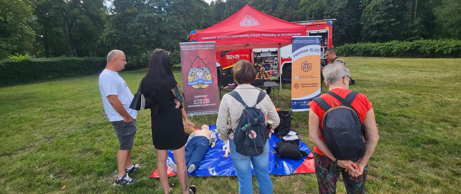 Zdjęcie pokazuje stanowisko do nauki pierwszej pomocy z perspektywy osób obserwujących szkolenie. Na pierwszym planie widać plecy i sylwetki uczestników zgromadzonych wokół rozłożonej na trawie niebieskiej plandeki z fantomami. W centrum uwagi trwa praktyczny instruktarz medyczny. W tle wyraźnie widać czerwony namiot Komendy Miejskiej Państwowej Straży Pożarnej w Wałbrzychu oraz banery promocyjne. Bezpośrednio za namiotem zaparkowany jest wóz bojowy.