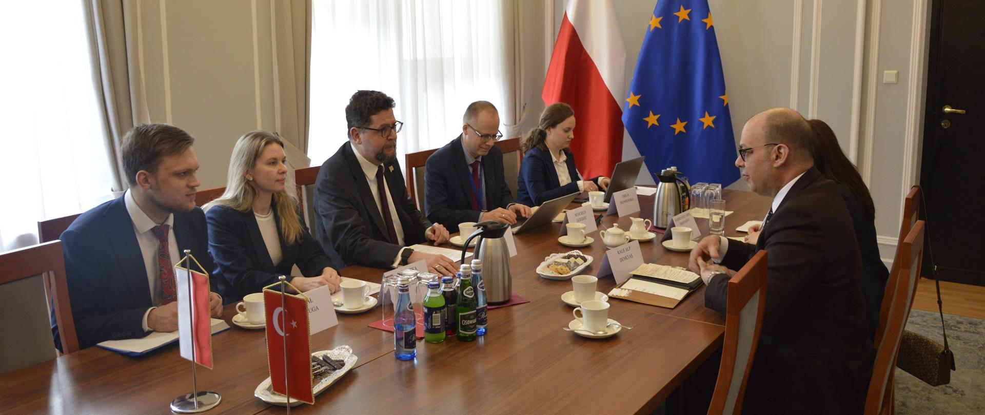 W małej sali przy podłużnym stole siedzi kilka rozmawiających osób, za stołem pod ścianą flagi Polski i UE.